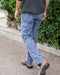 Person wearing blue pants and brown boots walking on a sidewalk with greenery in the background